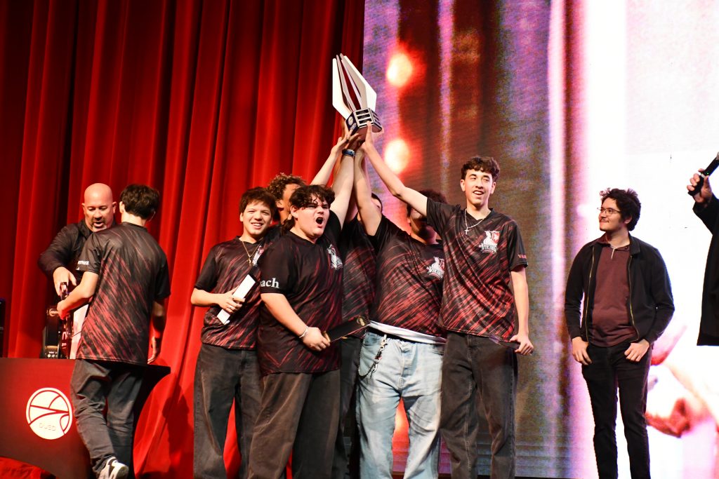Downey High School esports players celebrate on stage as they raise the championship trophy together, surrounded by teammates and coaches during the Downey Unified Esports Championship.