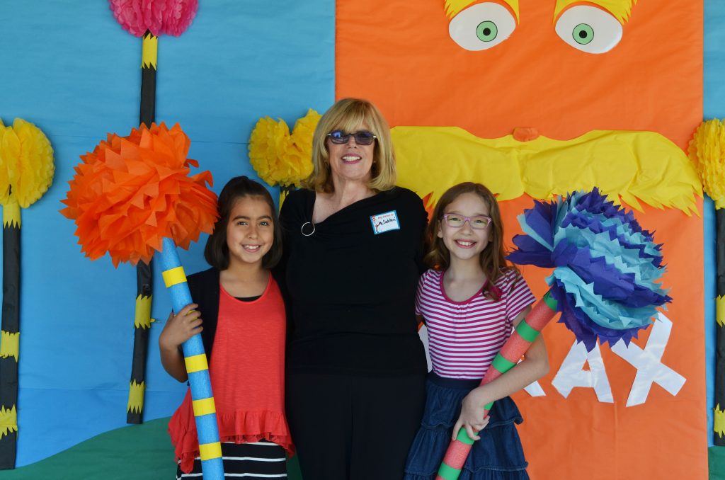 Former Downey Unified Board of Education Member Martha Sodetani stands with two elementary-age students in front of a colorful school event display decorated with oversized paper flowers and bright artwork.
