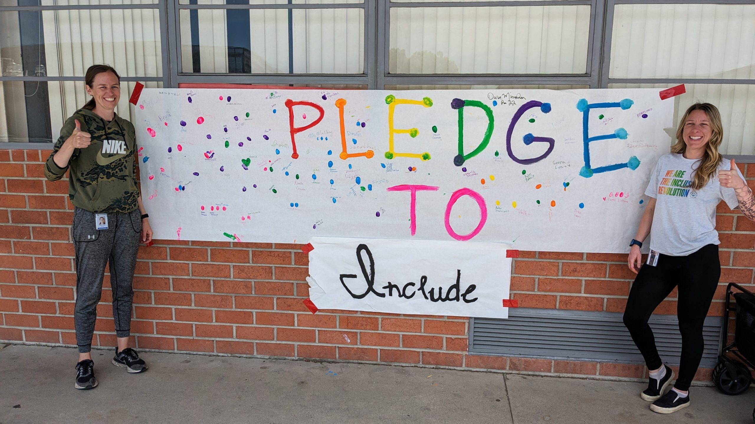 Two staff members from Doty Middle Schools stand beside a student-created “Pledge to Include” banner covered with colorful fingerprints and signatures.